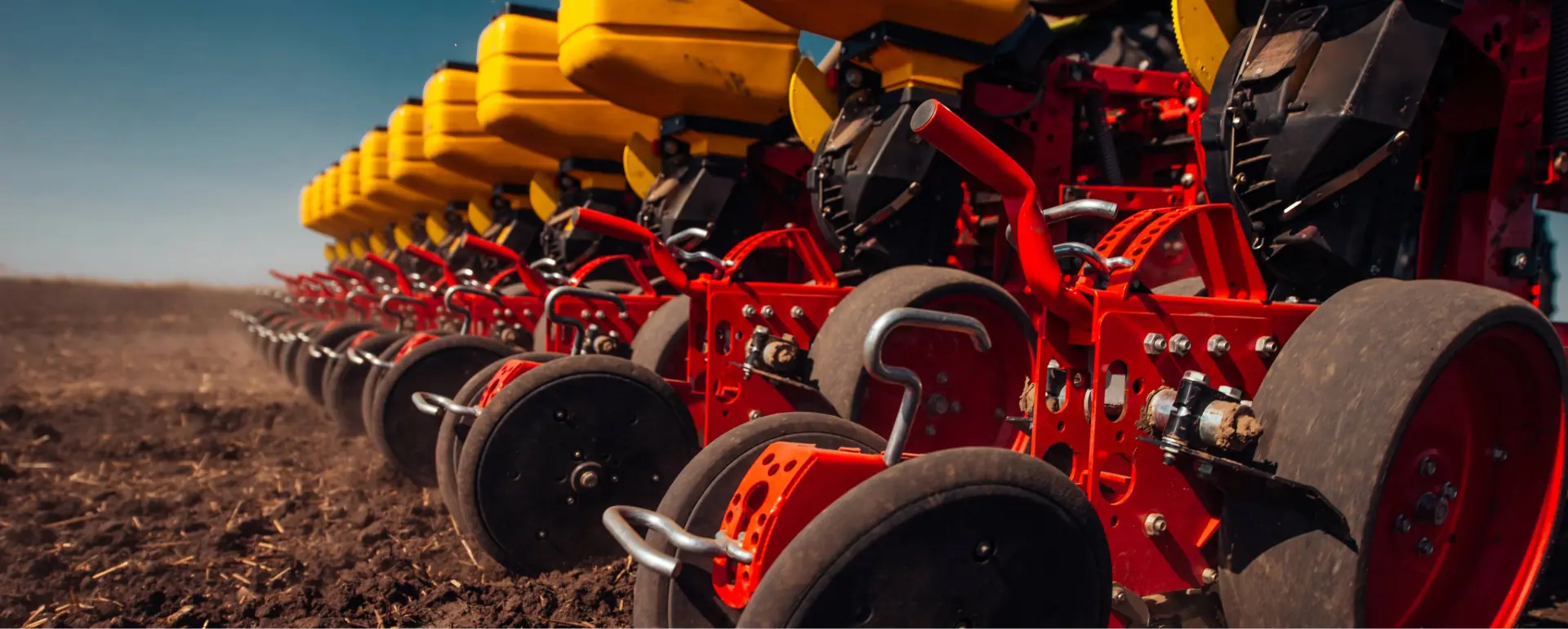 Vista em perspectiva das linhas de uma semeadora agrícola moderna nas cores vermelha e amarela, destacando os conjuntos de discos sulcadores e rodas compactadoras de borracha em contacto com o solo escuro.